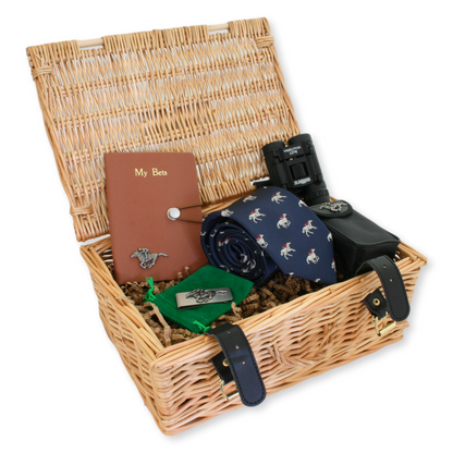 A wicker gift basket containing a notebook with a horse racing motif, binoculars, a navy blue tie with a racing horse and jockey design, and a stainless steel money clip.